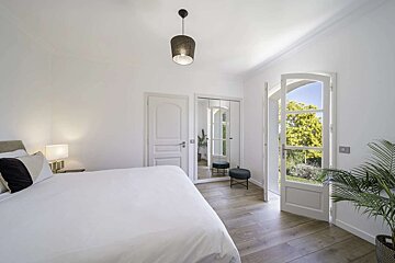 Bright, minimalist white bedroom with a large bed, open doors to a green garden, a mirrored closet, and light wood floors.