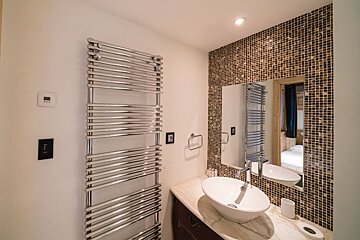 Modern bathroom with a chrome towel warmer, vessel sink vanity, and a mirror against a brown mosaic tiled wall, reflecting a bedroom.