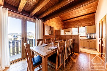 A cozy, rustic chalet interior features a wooden dining table, open kitchen, and a balcony with views of mountains and village houses, all with wood paneling.