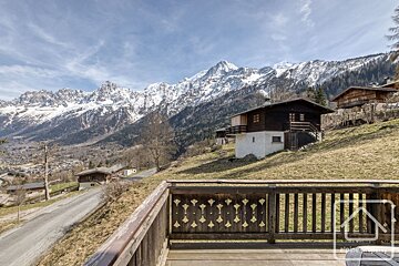 View from a wooden balcony overlooking traditional chalets and a valley, backed by impressive snow-capped mountains under a clear sky.