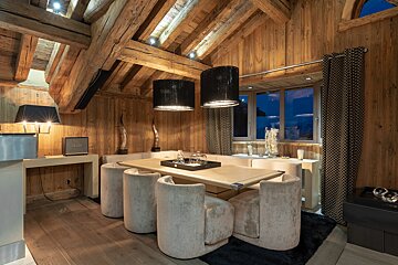 Luxurious chalet dining room with rich wooden beams/walls, plush seating, modern pendants, and a window looking out to a nighttime landscape.