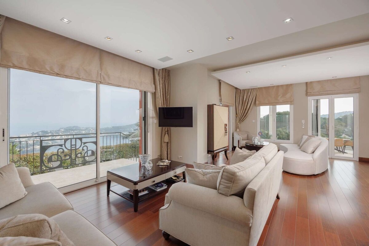 Modern living room with cream sofas, wood floors, and floor-to-ceiling windows offering a panoramic ocean and coastal town view.