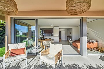 Sunny patio chairs looking into a modern open-plan living room and kitchen through large glass doors, with outdoor garden view.