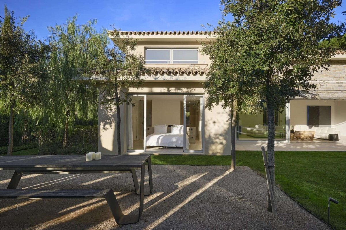 A modern villa with a visible bedroom through glass doors, an outdoor dining table on gravel, and lush trees under a clear blue sky.