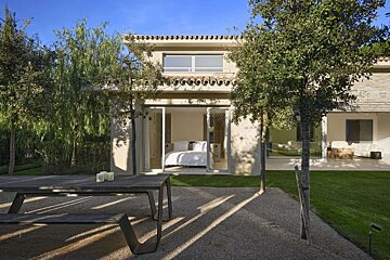 A modern villa with a visible bedroom through glass doors, an outdoor dining table on gravel, and lush trees under a clear blue sky.