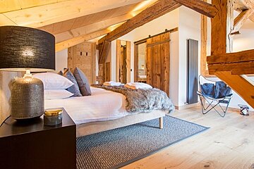 Rustic attic bedroom with exposed wood beams, a cozy bed with a fur throw, sliding barn doors, and a black bedside lamp.