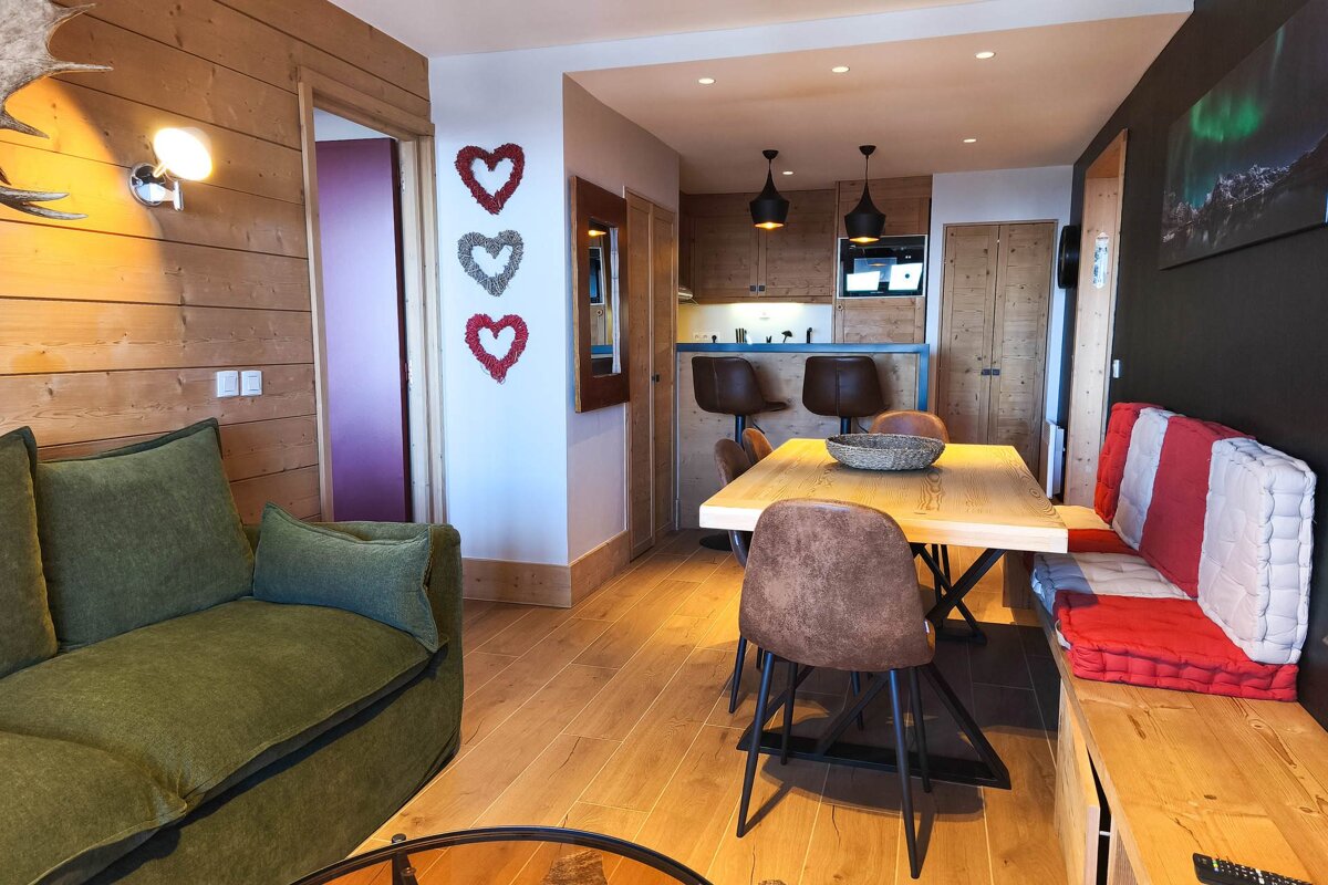 Cozy open-plan living space with wood accents, green sofa, dining table with bench, and kitchen. Features heart wall decor & Northern Lights art.
