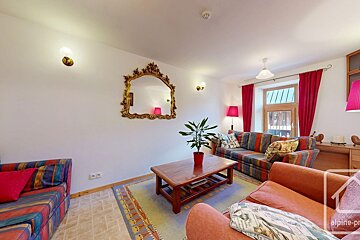 A bright living room with white walls, a gold mirror, red curtains, striped and orange sofas, a wooden coffee table, potted plant, and colorful rug.