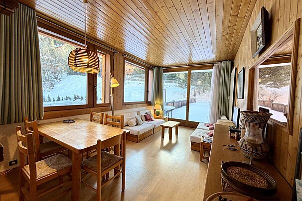 Cozy, wood-paneled cabin interior with dining area and sofas. Large windows overlook a snowy winter landscape, creating a warm atmosphere.