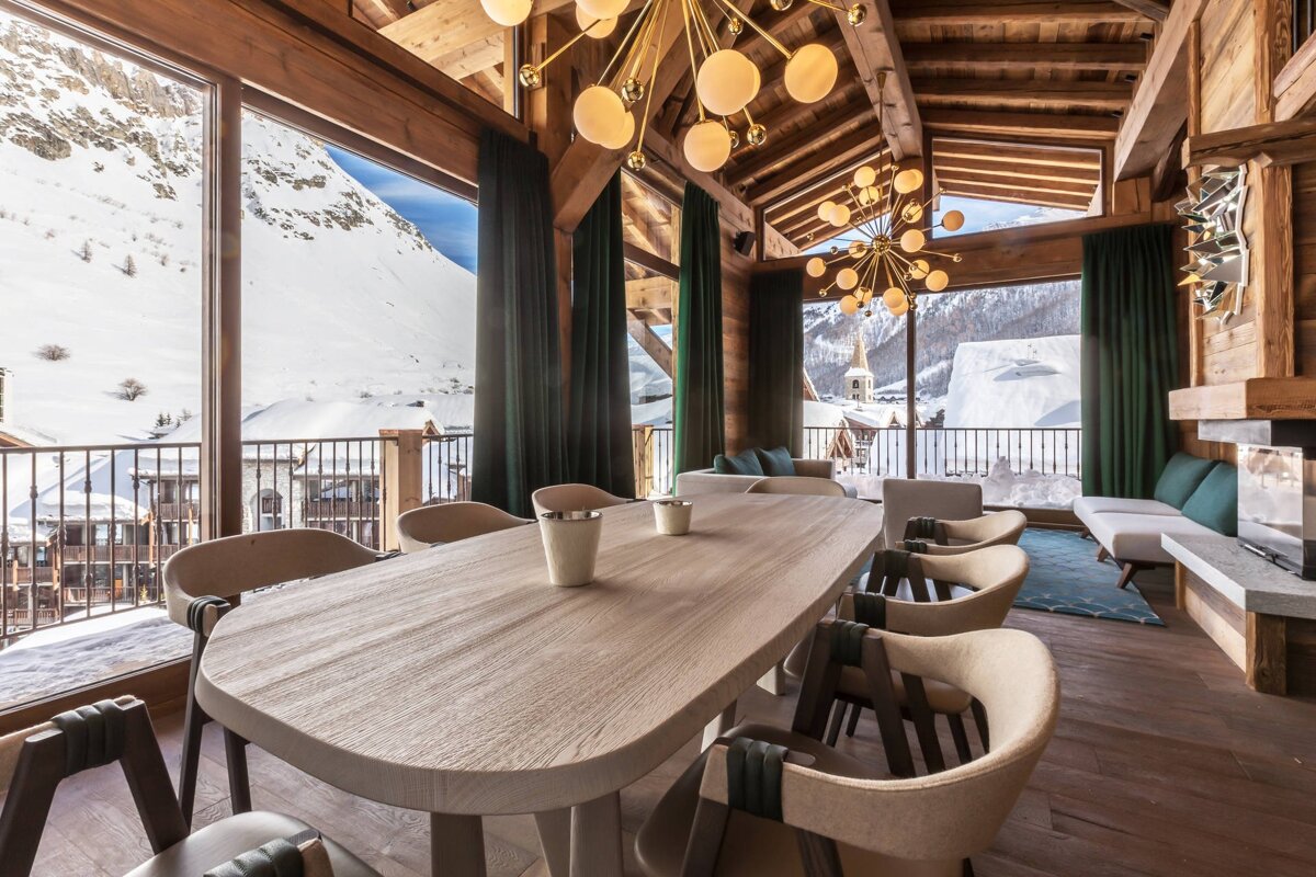 Luxurious ski chalet dining/living room with a long wooden table, modern chandeliers, and panoramic windows overlooking snow-covered mountains.