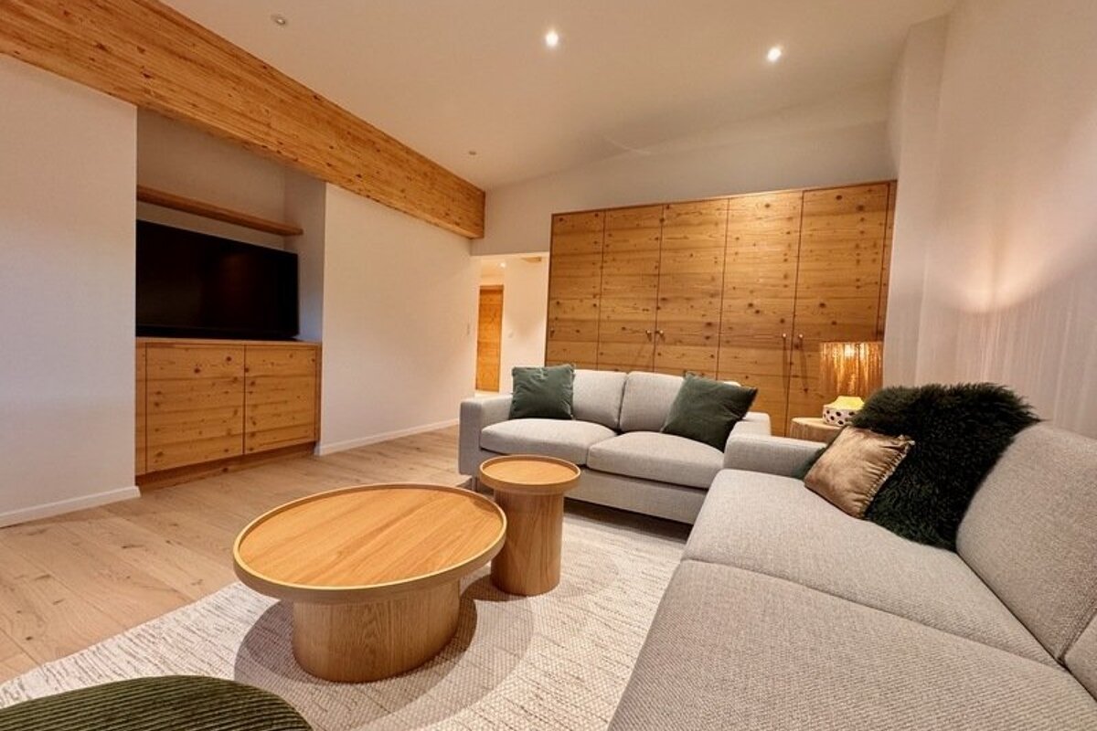 Modern, cozy living room featuring grey sofas, wooden coffee tables, wood-paneled walls, and warm lighting.