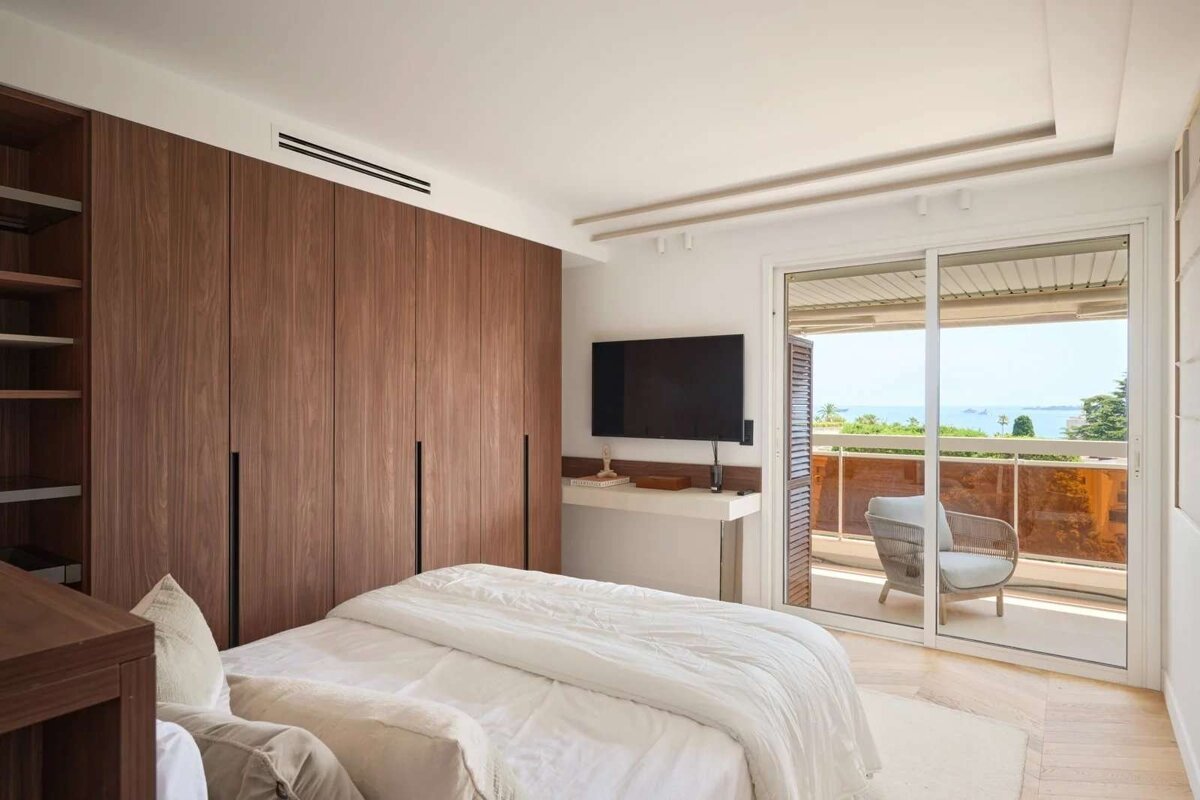 Modern bedroom with wooden wardrobe, bed, TV, and a balcony overlooking a scenic sea view.