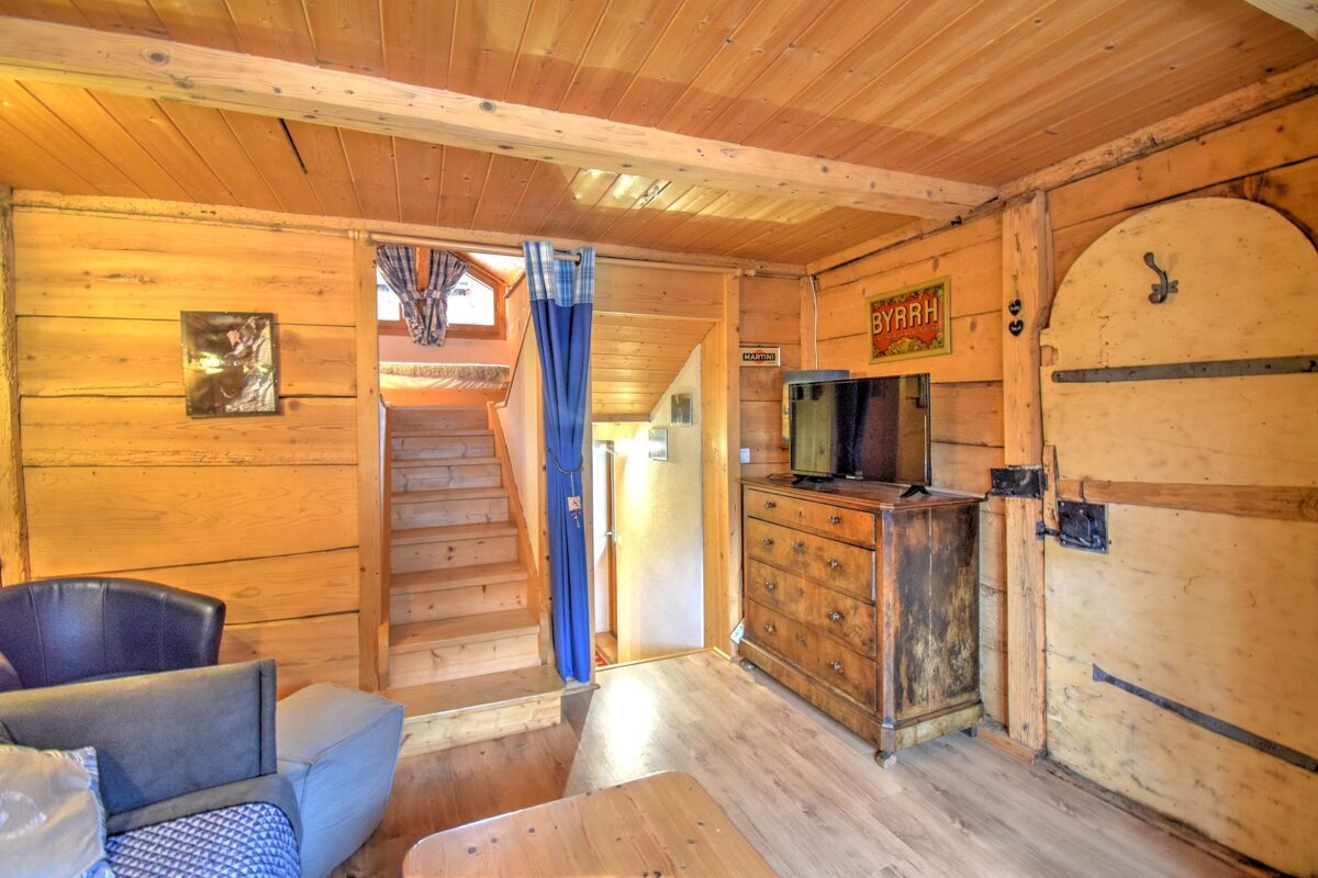 A cozy, rustic wooden room with stairs, a TV on an antique dresser, and a unique rounded door.