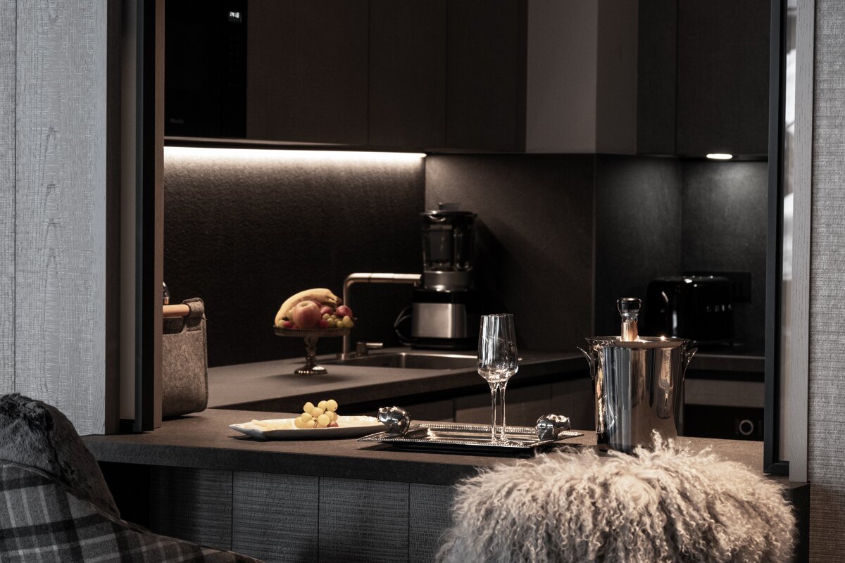 A sleek, dimly lit dark kitchen bar with a champagne bucket, wine glass, fruit, and grapes on the counter, illuminated by under-cabinet lighting.
