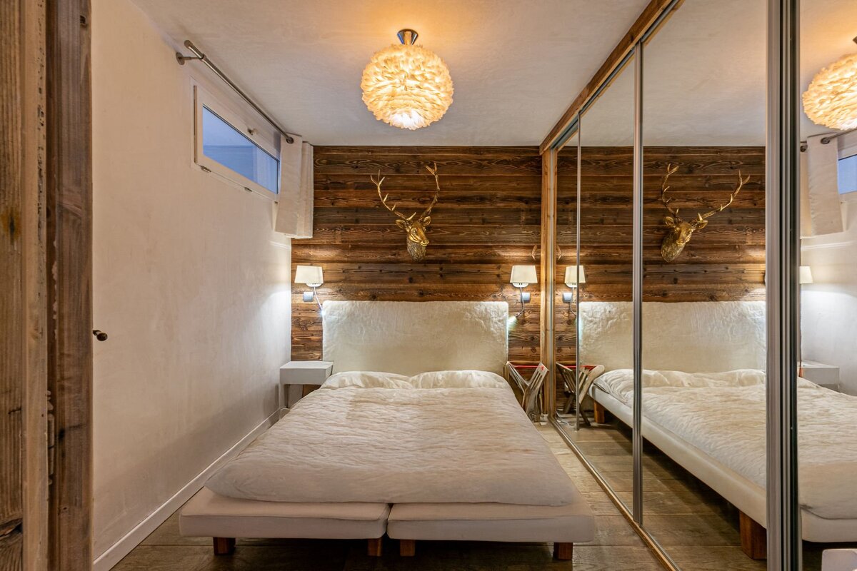 A cozy, rustic bedroom featuring a white bed, a wooden accent wall with a golden deer head, mirrored closet, and a unique feather-like light.