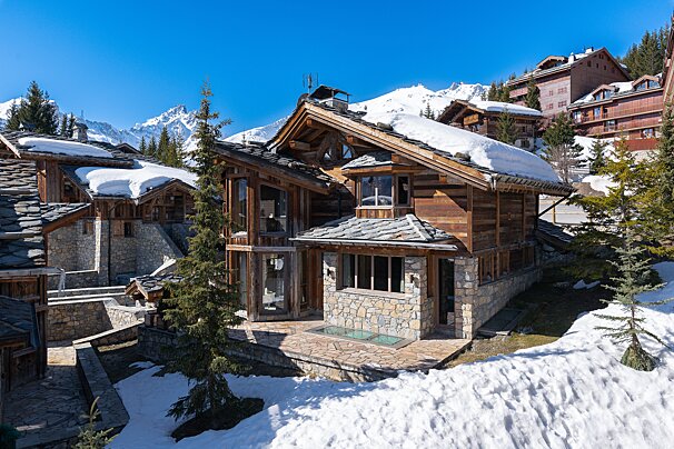 A beautiful wooden chalet with stone accents and snow-dusted roofs in a sunny mountain village, surrounded by snow and distant peaks.
