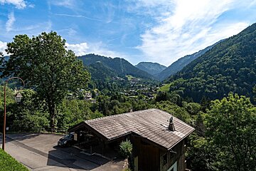 Scenic view of a lush green mountain valley with a village, traditional building, and car in the foreground under a blue, cloudy sky.