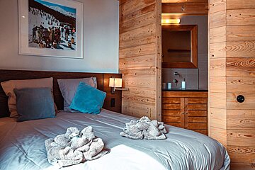 A cozy bedroom with light blue bedding, decorative towels, and a ski-themed print. Rustic wooden walls blend into an integrated bathroom with a sink.