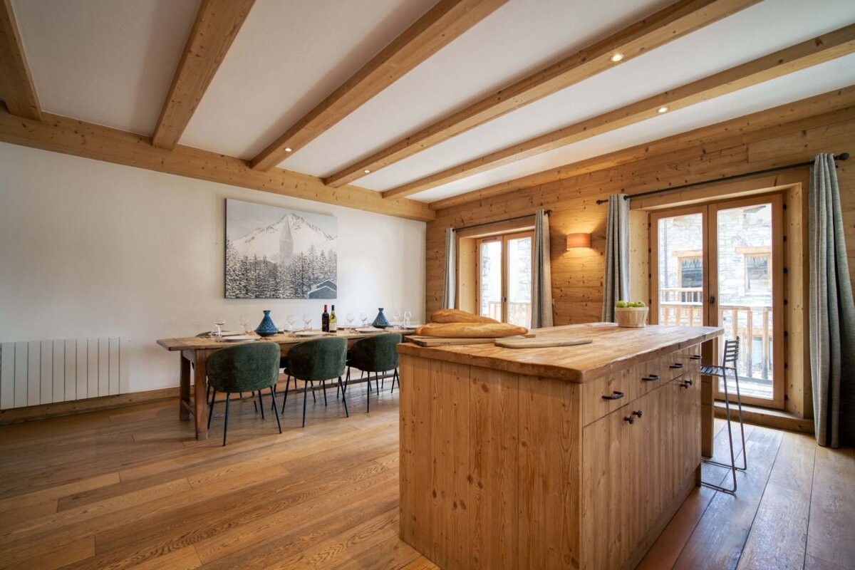 Rustic, chalet-style dining and kitchen area with wooden beams, floors, walls, a long dining table, island, mountain art, and balcony doors.