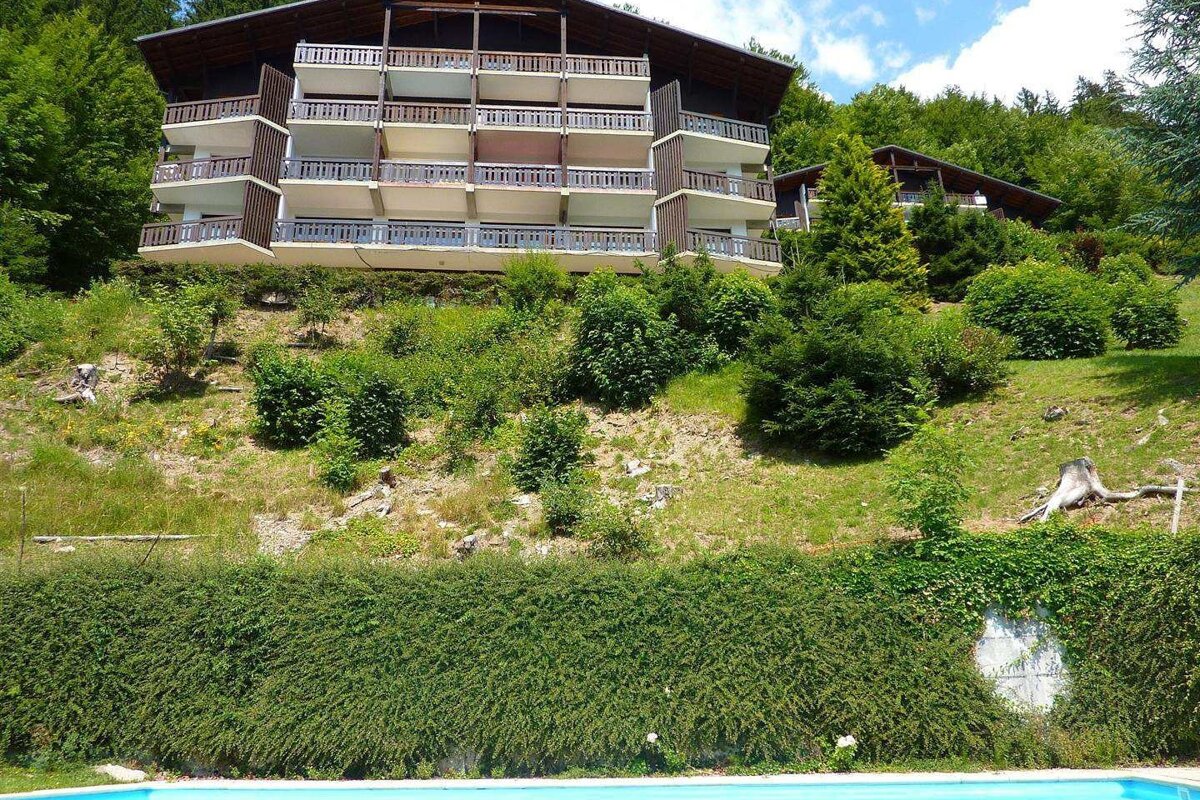 Multi-story apartment buildings on a lush green hillside with trees and shrubs, overlooking a bright blue swimming pool in the foreground.