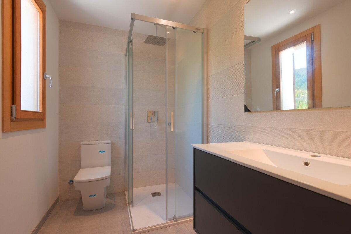 A bright, modern bathroom featuring a glass shower, toilet, dark vanity with white sink, mirror, and a wooden-framed window.