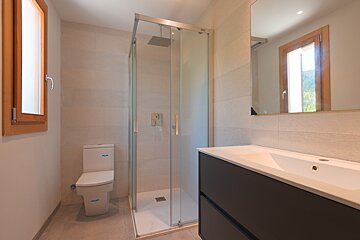 A modern bathroom featuring a toilet, a walk-in glass shower, a dark vanity with an integrated sink, a large mirror, and a wood-framed window.