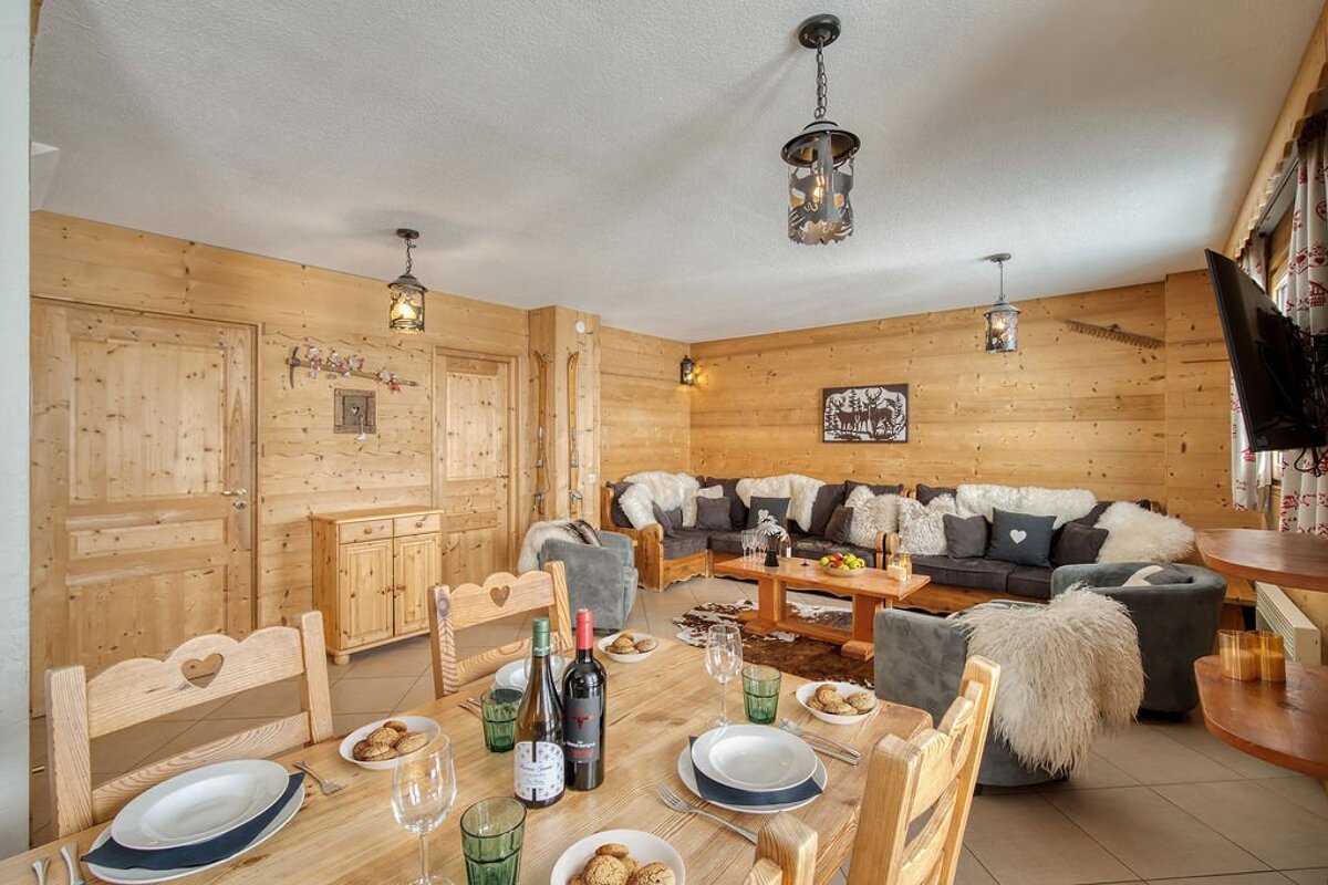 A warm, rustic chalet living and dining room featuring light wood paneling, a dining table set with wine, and a cozy lounge area with sofas and fur throws.