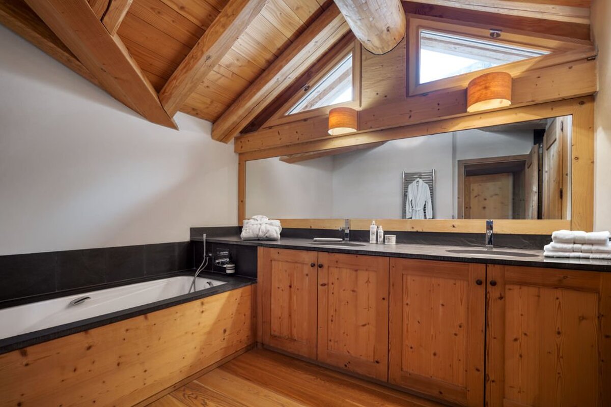 Modern rustic bathroom with exposed wooden beams, a built-in tub, double vanity with large mirror, and angled windows.