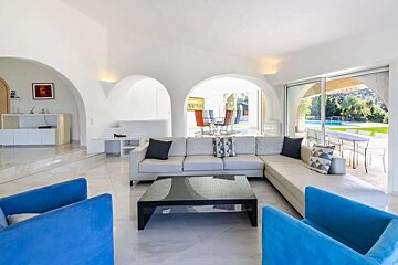 A modern, white living room with arched doorways, a large L-shaped sofa, blue armchairs, and a black coffee table, opening to an outdoor pool.