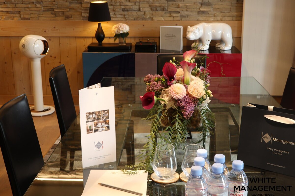 A modern meeting setup features a glass table with a vibrant floral centerpiece, water bottles, glasses, and a brochure. A white bear statue and lamp decorate the background.