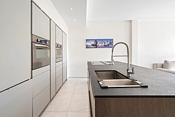 A modern kitchen with white built-in ovens, a dark island featuring an integrated sink and pull-down faucet, and a tiled floor.
