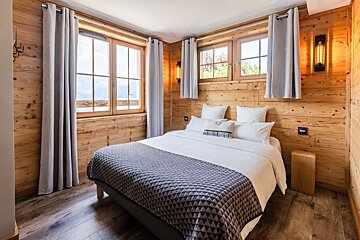 Cozy cabin bedroom with wood-paneled walls, a neat bed with a grey patterned blanket, and windows framing mountain views.