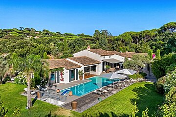 An aerial view of a luxurious white villa with a blue pool, sun loungers, and green gardens, nestled in a forested hillside under a clear blue sky.