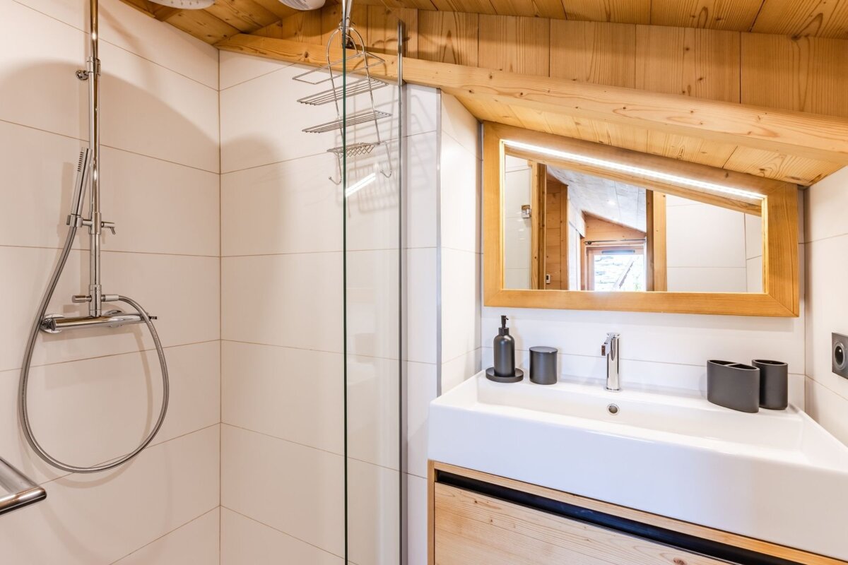 Cozy rustic bathroom featuring a glass shower, white sink, and an illuminated mirror under a sloped wooden ceiling.