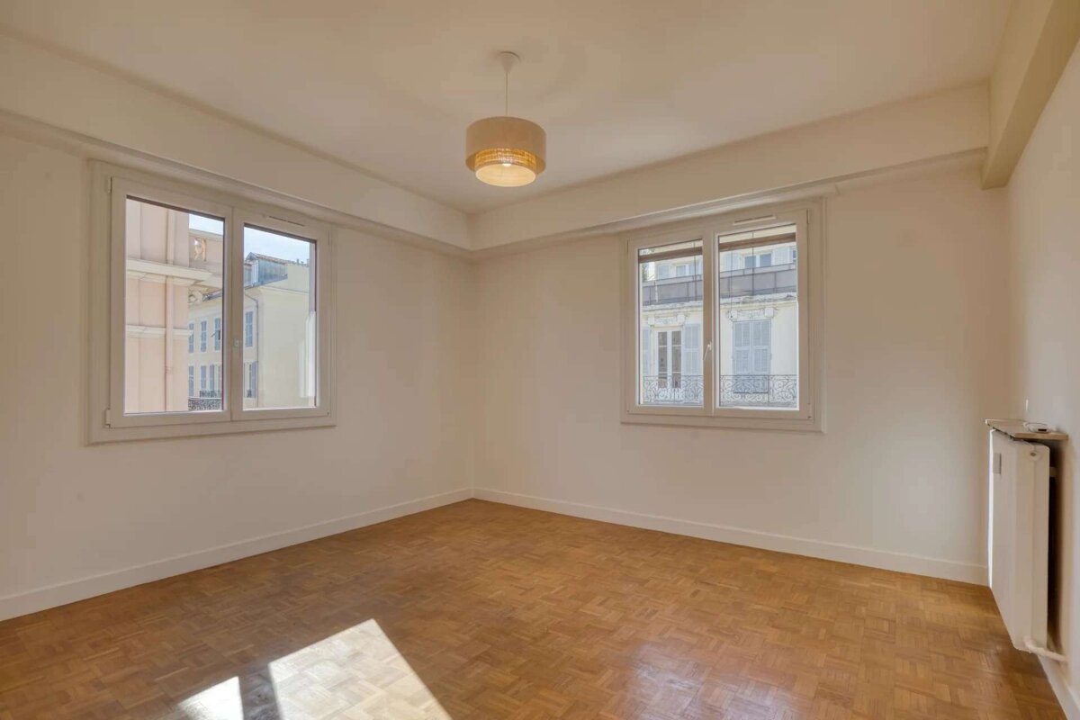 Bright, empty room with white walls, parquet floor, two windows showing city views, and a ceiling light. Sunlight illuminates the space.