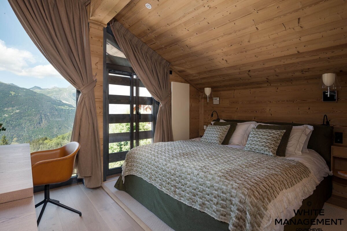 Wooden chalet bedroom with a large bed, a leather chair, and large windows revealing a scenic mountain view.