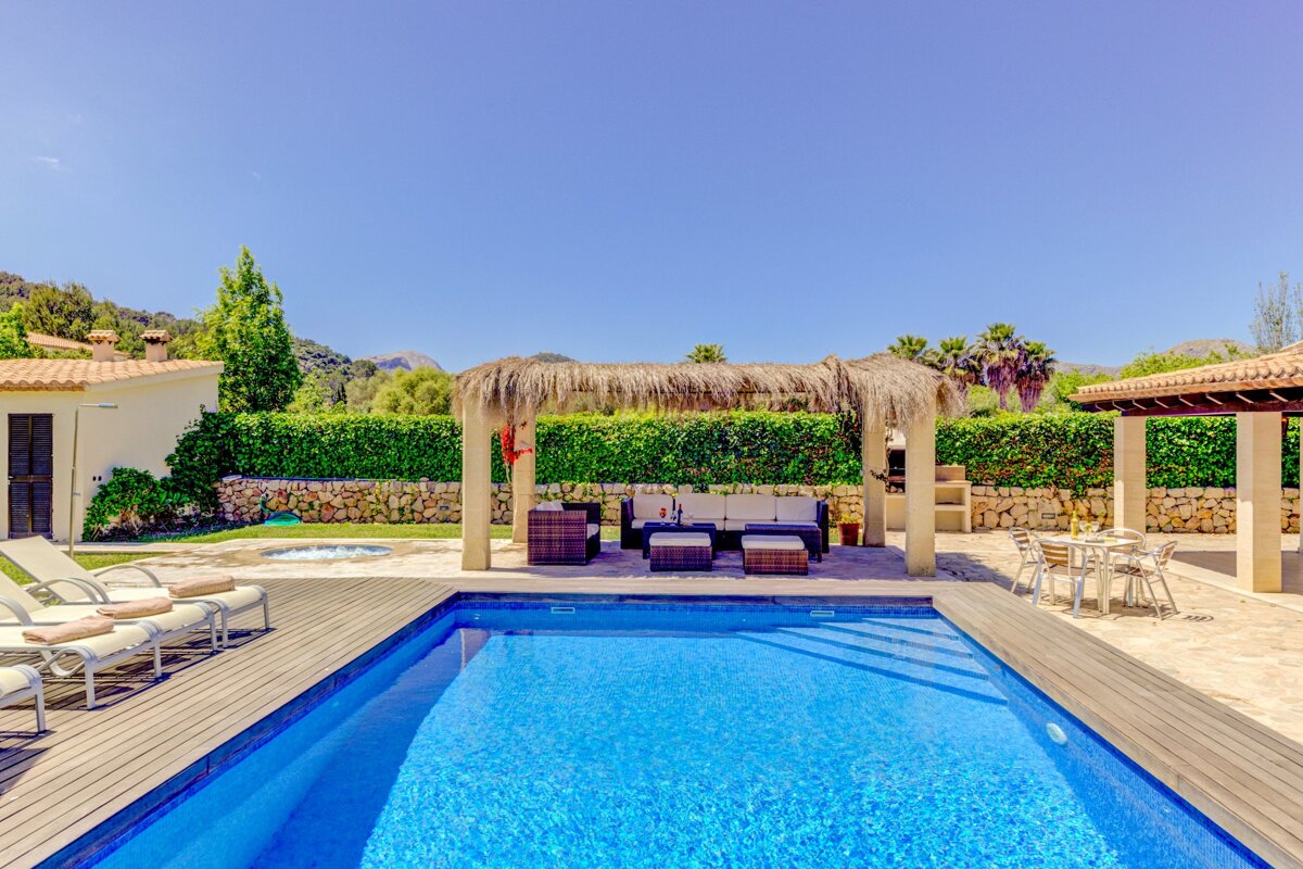 A sunny villa backyard features a sparkling blue swimming pool, a thatched pergola with outdoor seating, sun loungers, and a dining area, all under a clear sky.