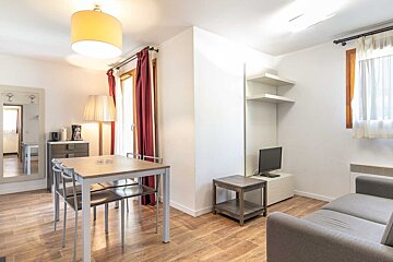 A bright, modern living/dining room with a wooden table, gray sofa, TV, and large window with red curtains, all on a wooden floor.