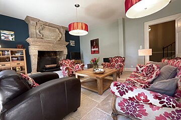 A cozy, eclectic living room with a large stone fireplace, vibrant red floral seating, dark leather chairs, and two red pendant lights.