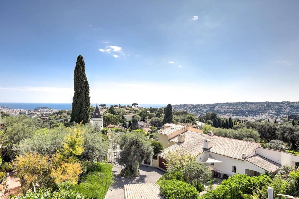 A vast elevated view of a vibrant coastal landscape with white houses, red roofs, lush trees, and a tall cypress, all set against a clear blue sky and sea.