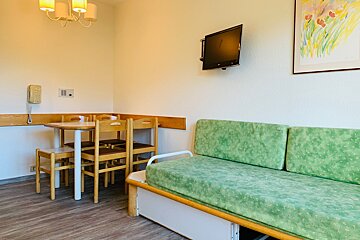 A bright room features a green sofa, wooden dining table with chairs, a wall-mounted TV, telephone, and colorful artwork.