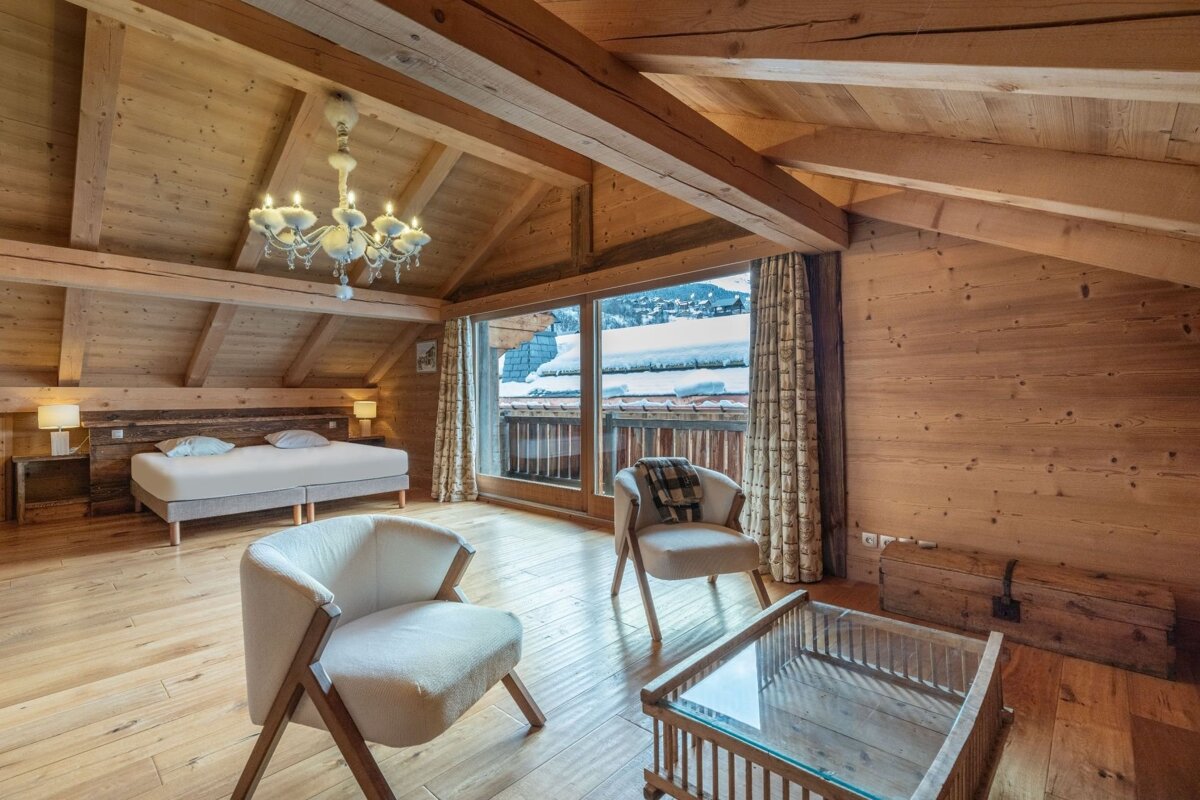 Warm, all-wood chalet room with a bed, seating area, and large window offering snowy mountain views. Exposed beams and a chandelier add character.