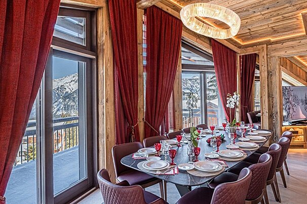 A luxurious alpine chalet dining room with a long table set for dinner, deep red curtains, and large windows revealing a stunning snowy mountain view.