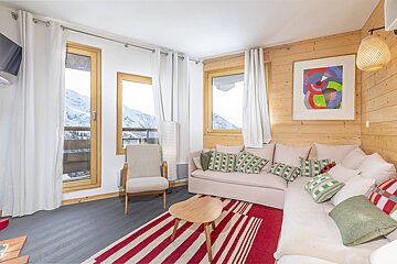 Cozy chalet living room with snowy mountain views, featuring a cream sectional sofa, wood paneling, and a red striped rug.