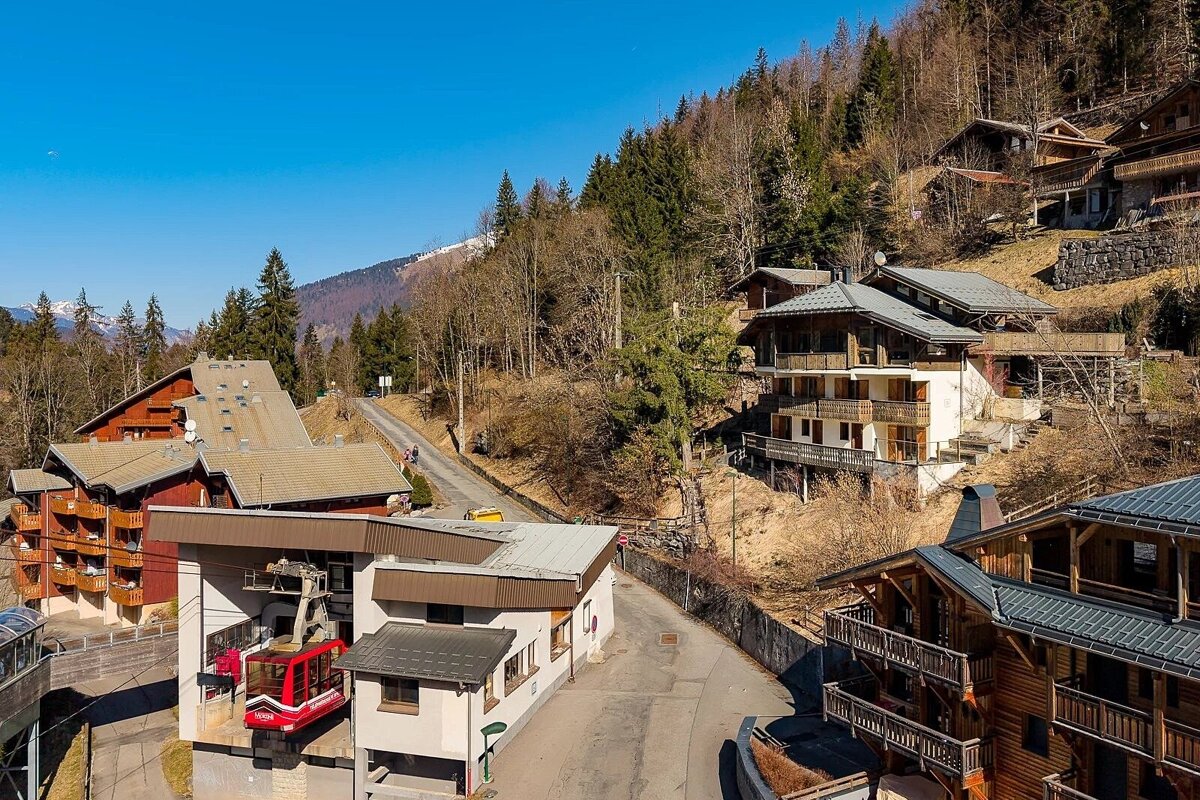 Alpine village with wooden chalets, a red cable car station, and forested hills under a bright blue sky.