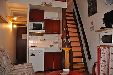 A compact studio apartment with a kitchenette, sofa, and a wooden staircase leading to a loft bed. Warm lighting creates a cozy, practical living space.