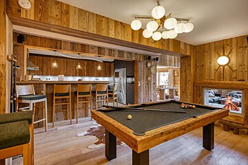 A cozy, wood-paneled room with a billiards table, modern kitchen/bar area, and a glass-enclosed fireplace, exuding a rustic yet chic vibe.
