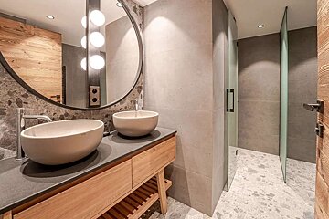 A modern bathroom features a double wood vanity with vessel sinks, a large round mirror with integrated lights, and a glass-partitioned shower. Grey stone and wood accents create a contemporary look.