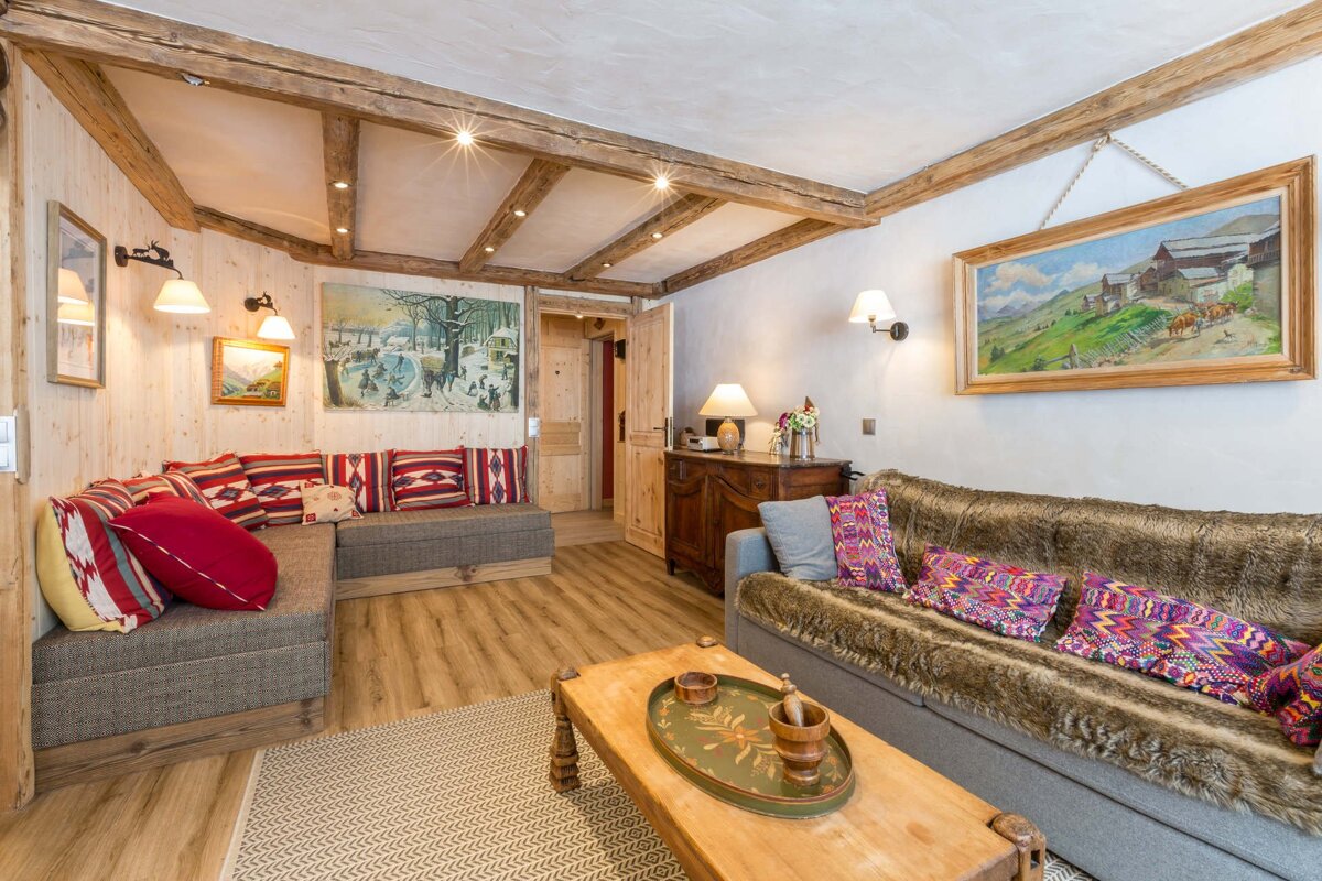 A cozy, rustic chalet living room with wooden beams, light wood paneling, upholstered benches, sofas, and art, creating a warm ambiance.