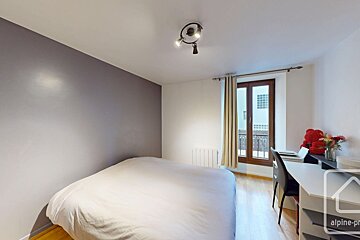 A bedroom with a grey accent wall, white bed, ceiling light, wooden floors, and a study desk by a glass door.